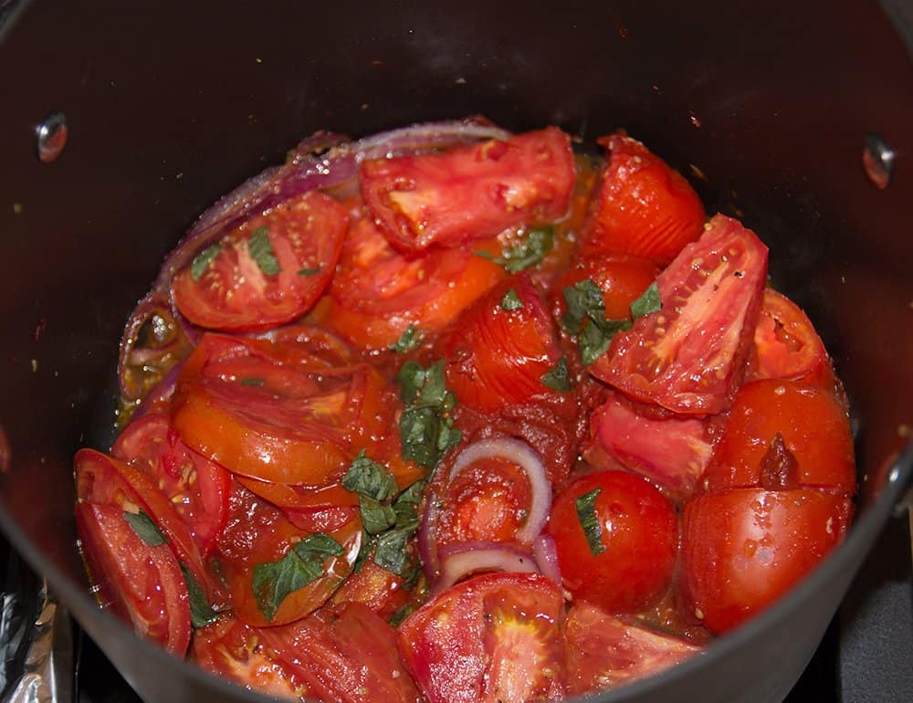 The tomato sauce simmering on the stove. Use a spoon to break the tomatoes apart in order to smooth out the sauce.